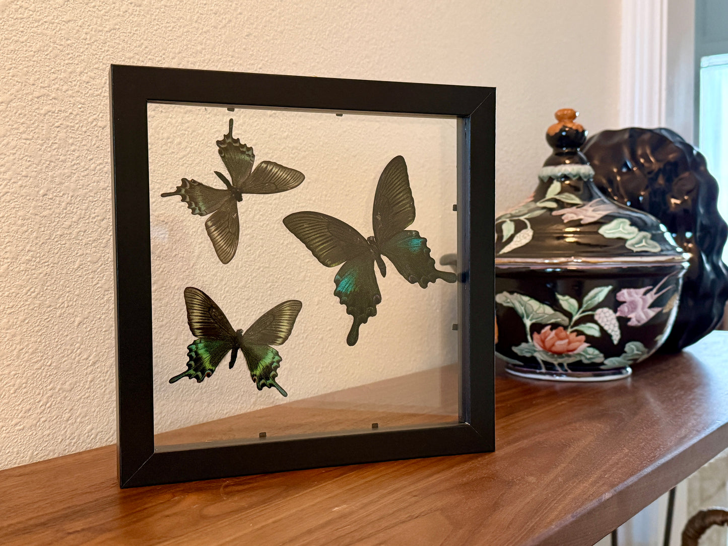 Framed butterfly artwork on a wooden surface with a decorative pot in the background.