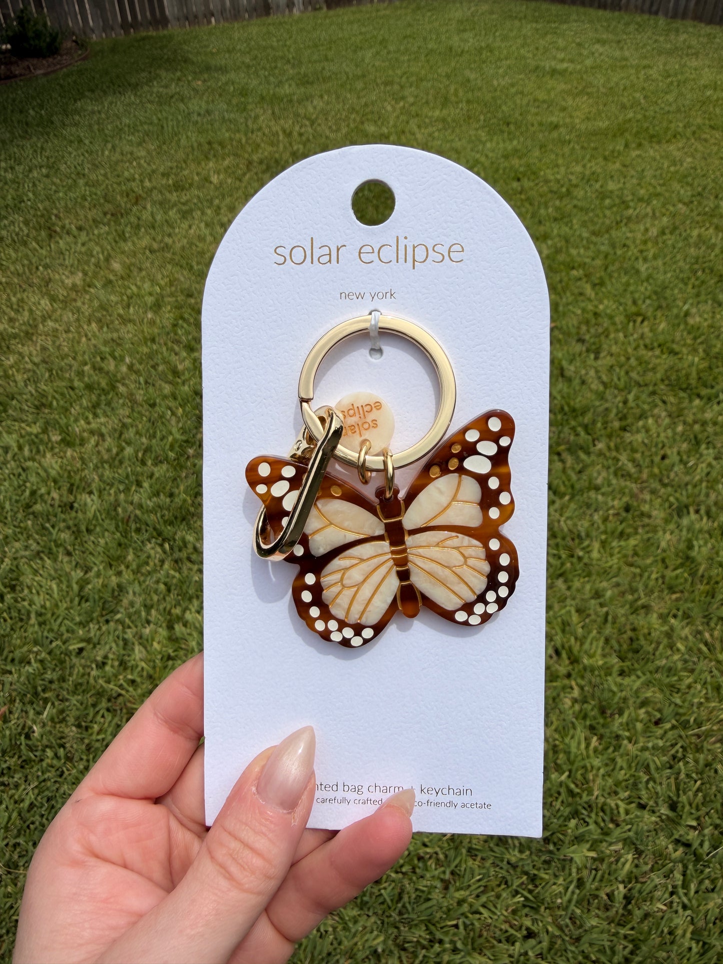 A butterfly-shaped solar keychain on a white card labeled 'solar eclipse,' held by a hand against a green grass background.